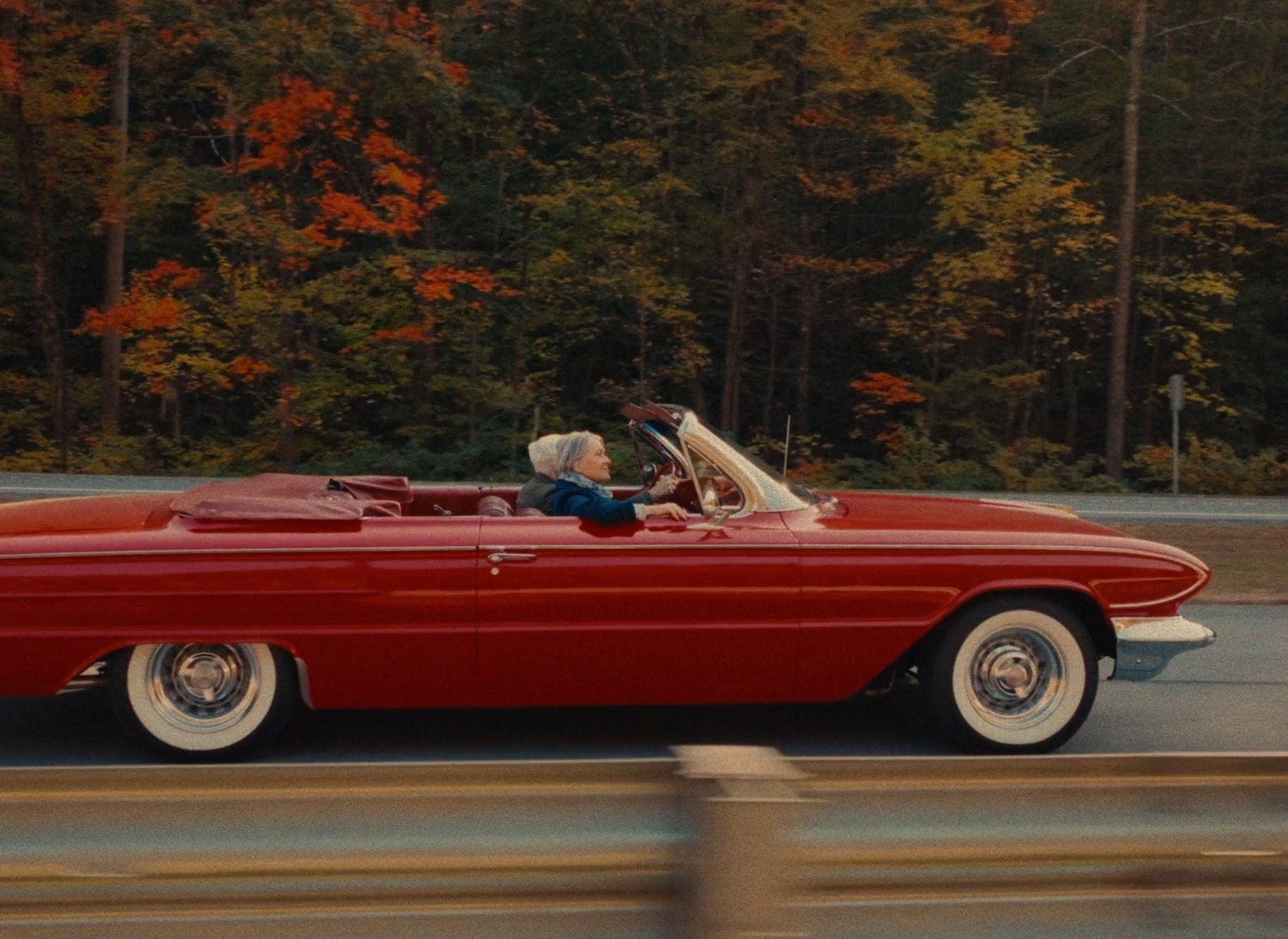 Red convertible tracking past a fall forest, driver at the wheel