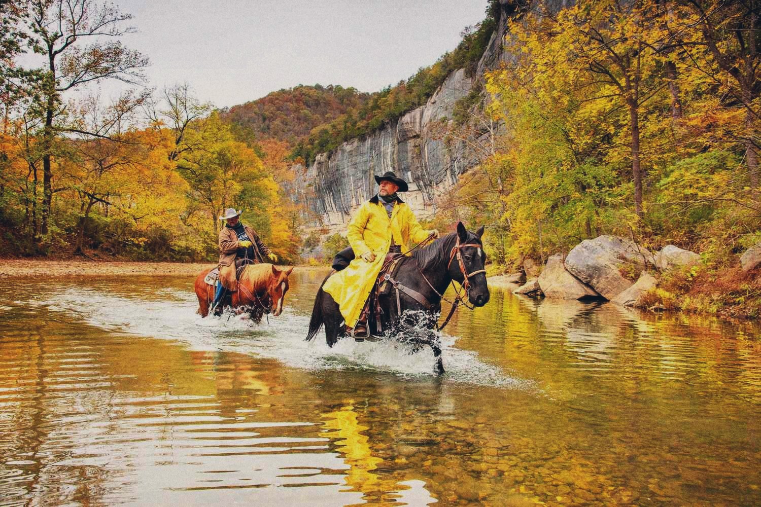 Two riders on horseback crossing a shallow river below a tall bluff, with trees in yellow fall color