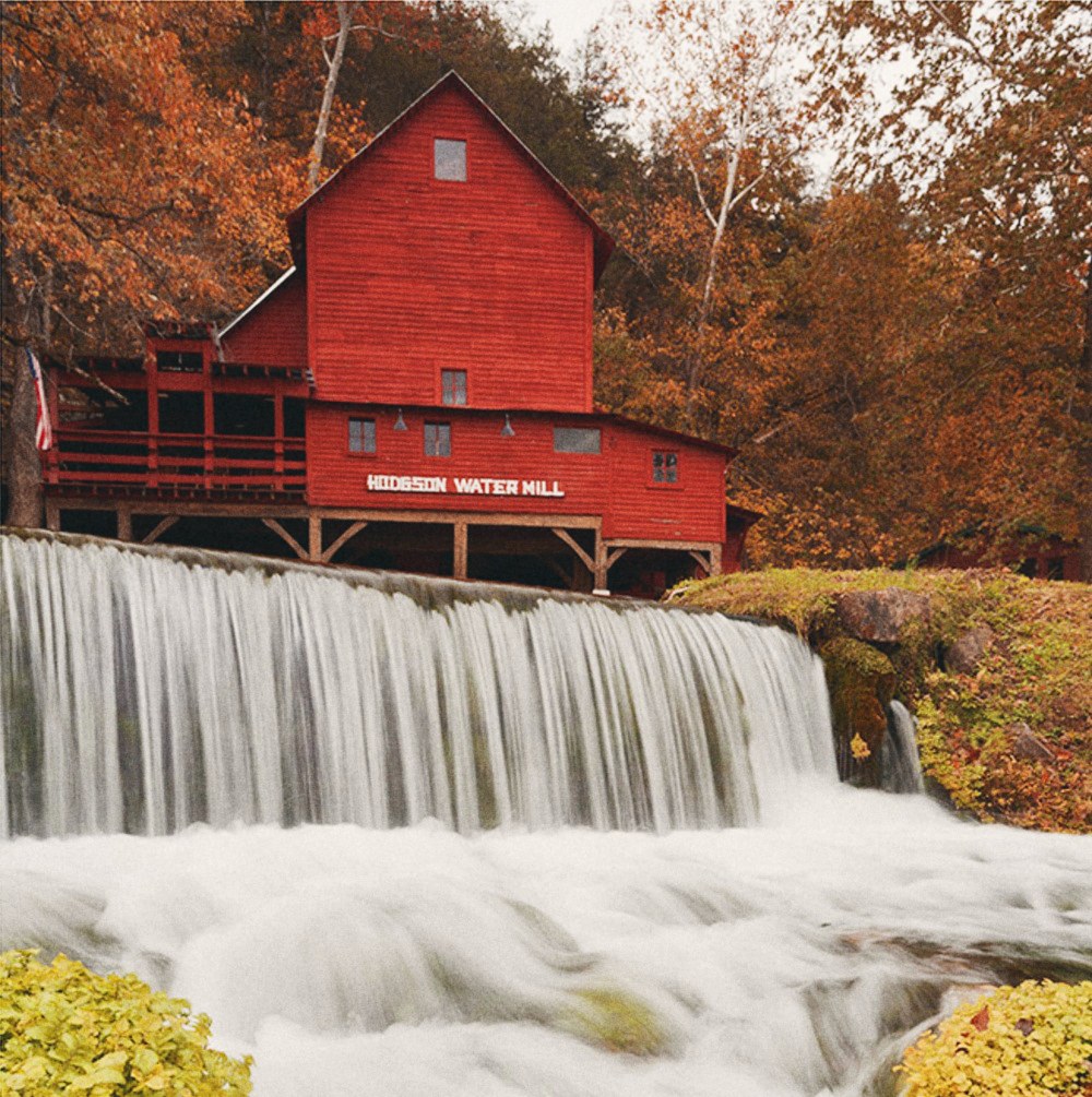 A red wooden water mill above a wide waterfall, surrounded by trees