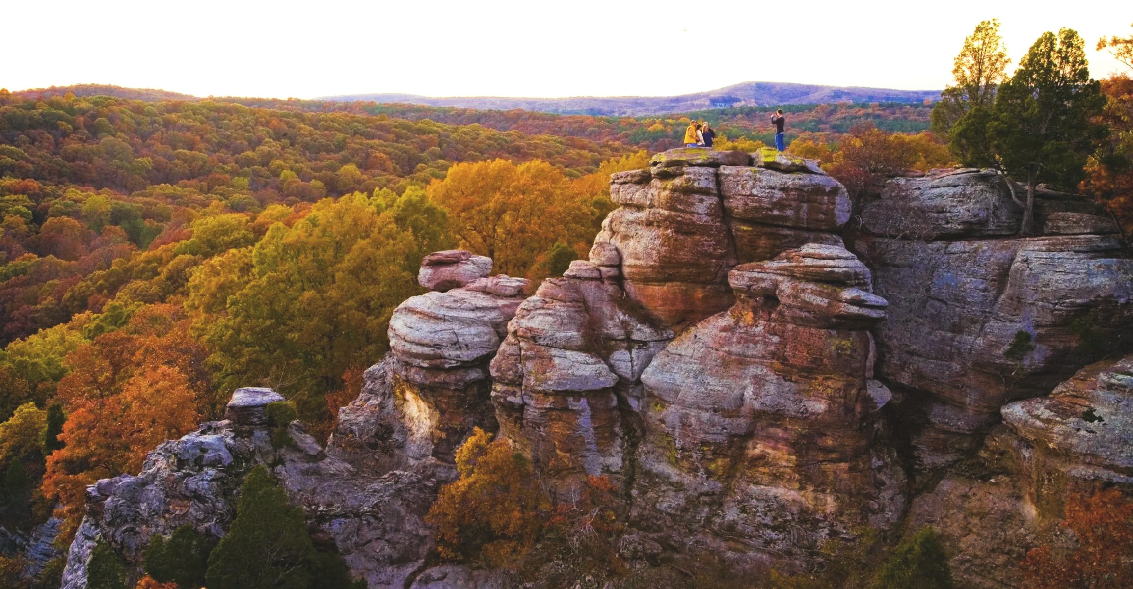Travelers on the sandstone bluffs of Garden of the Gods above a forest in fall color at sunset